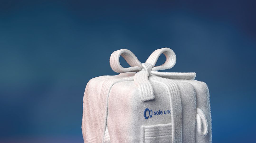 Weihnachtlich verpacktes Wellness-Geschenk mit dem Logo von sole uno auf einer weichen, weißen Frottier-Tasche vor blau-violettem Hintergrund.