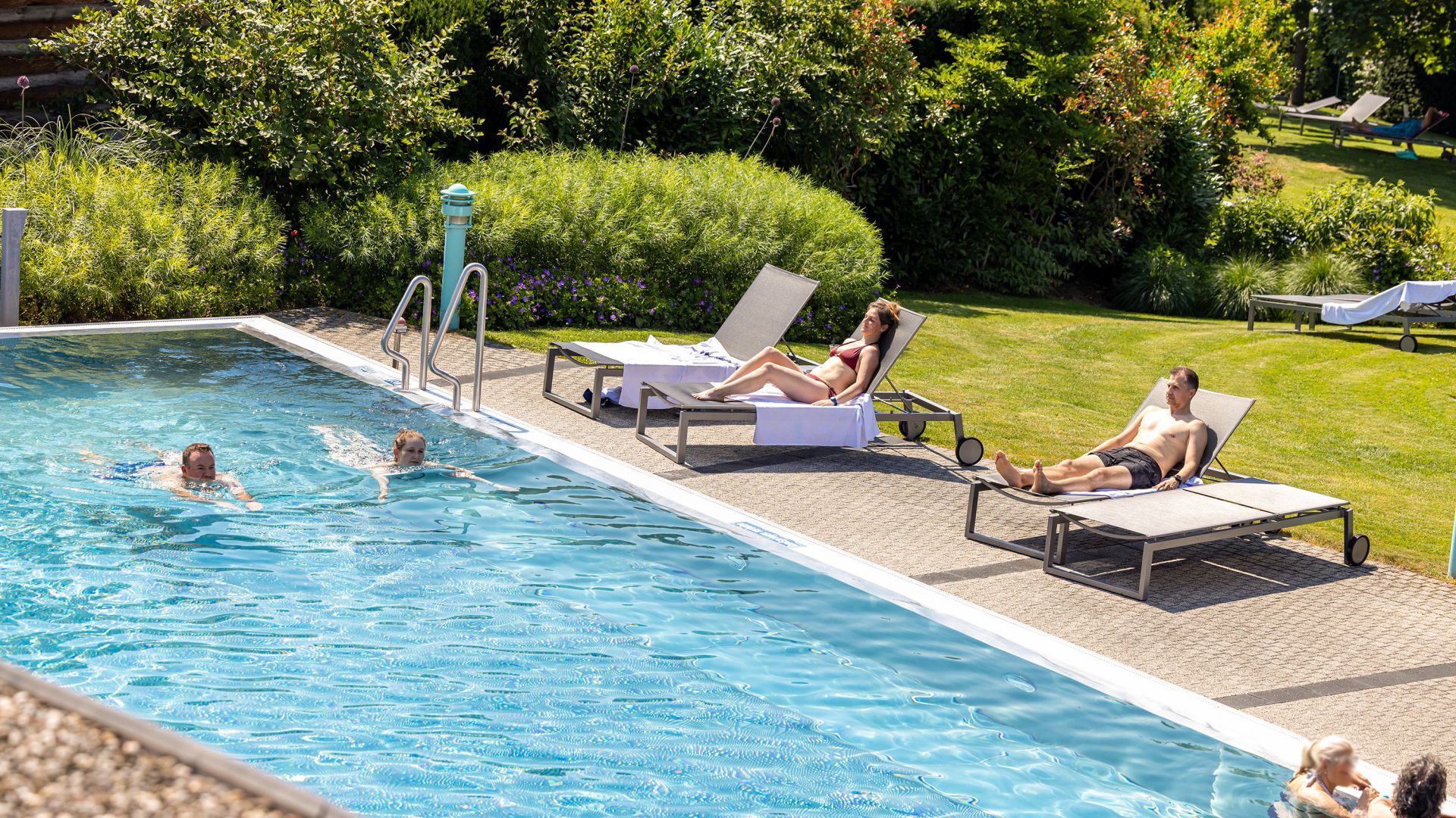 Badegäste beim entspannten Schwimmen im türkisblauen Aussenbecken des sole uno Rheinfelden, flankiert von Sonnenanbetern auf Liegen in einer grünen, gepflegten Parkanlage.