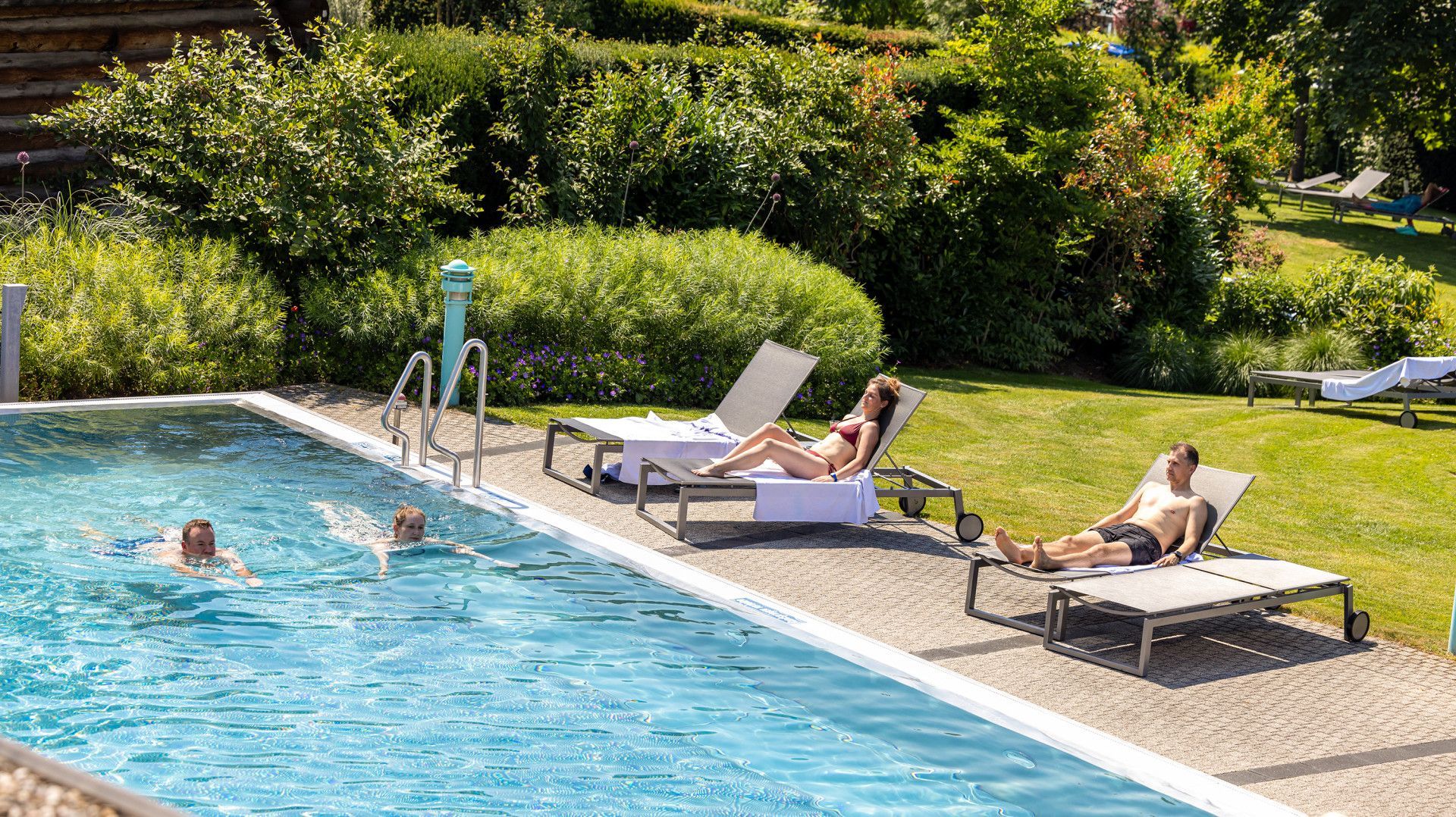 Badegäste beim entspannten Schwimmen im türkisblauen Aussenbecken des sole uno Rheinfelden, flankiert von Sonnenanbetern auf Liegen in einer grünen, gepflegten Parkanlage.