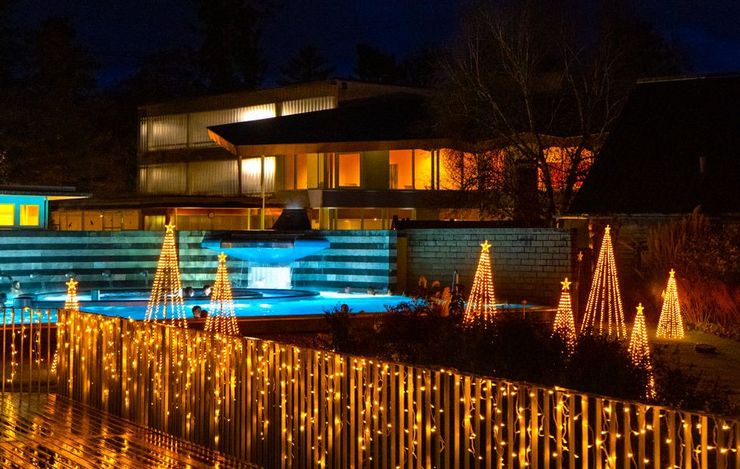 Festlich beleuchtete Thermalbadanlage bei Nacht mit blauem Poollicht und goldenen Lichterketten im Vordergrund.