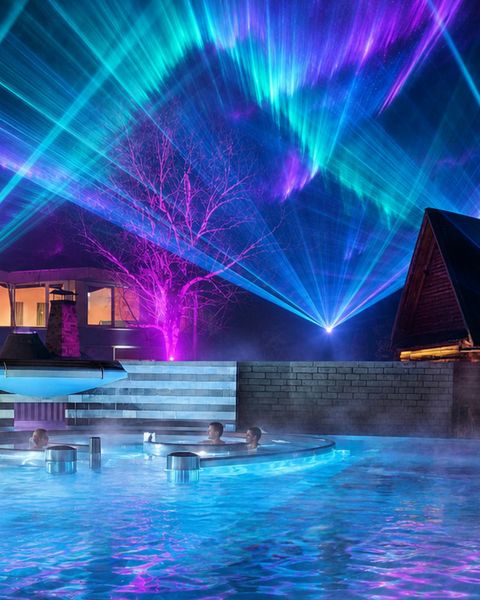 Gäste im dampfenden Außenbecken der Wellness-Welt sole uno in Rheinfelden während einer bunten Lichtkunstshow am Nachthimmel.
